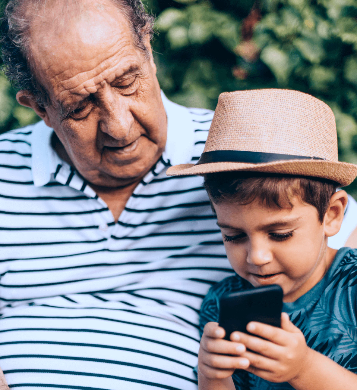 Homem idoso sorridente de camisa listrada observando carinhosamente seu neto de chapéu usando smartphone em ambiente ao ar livre