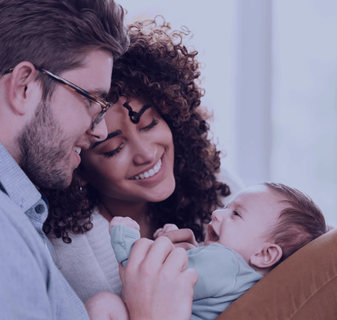 Casal sorridente segurando bebê recém-nascido, pai de óculos e barba ao lado da mãe de cabelos cacheados, todos em momento carinhoso de conexão familiar