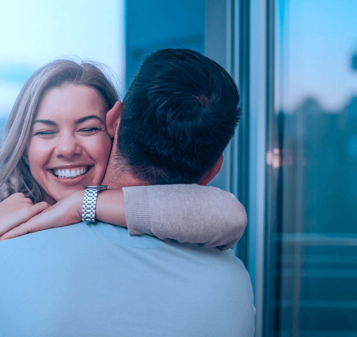 Casal sorridente se abraçando carinhosamente próximo a uma janela, mulher loira radiante olhando para a câmera enquanto abraça homem de cabelos escuros, representando confiança nos serviços Hemocord