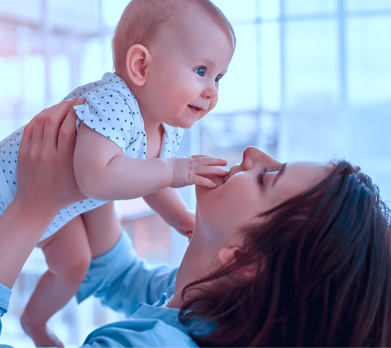 Mãe morena segurando bebê de olhos azuis vestindo body branco com estampa de estrelas, em momento íntimo de interação e brincadeira entre mãe e filho com luz natural suave ao fundo