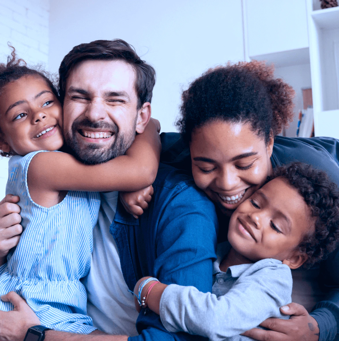 Família multirracial de quatro pessoas se abraçando carinhosamente em casa, com pai caucasiano, mãe negra e duas crianças de cabelos cacheados, todos sorrindo em momento de afeto e união familiar