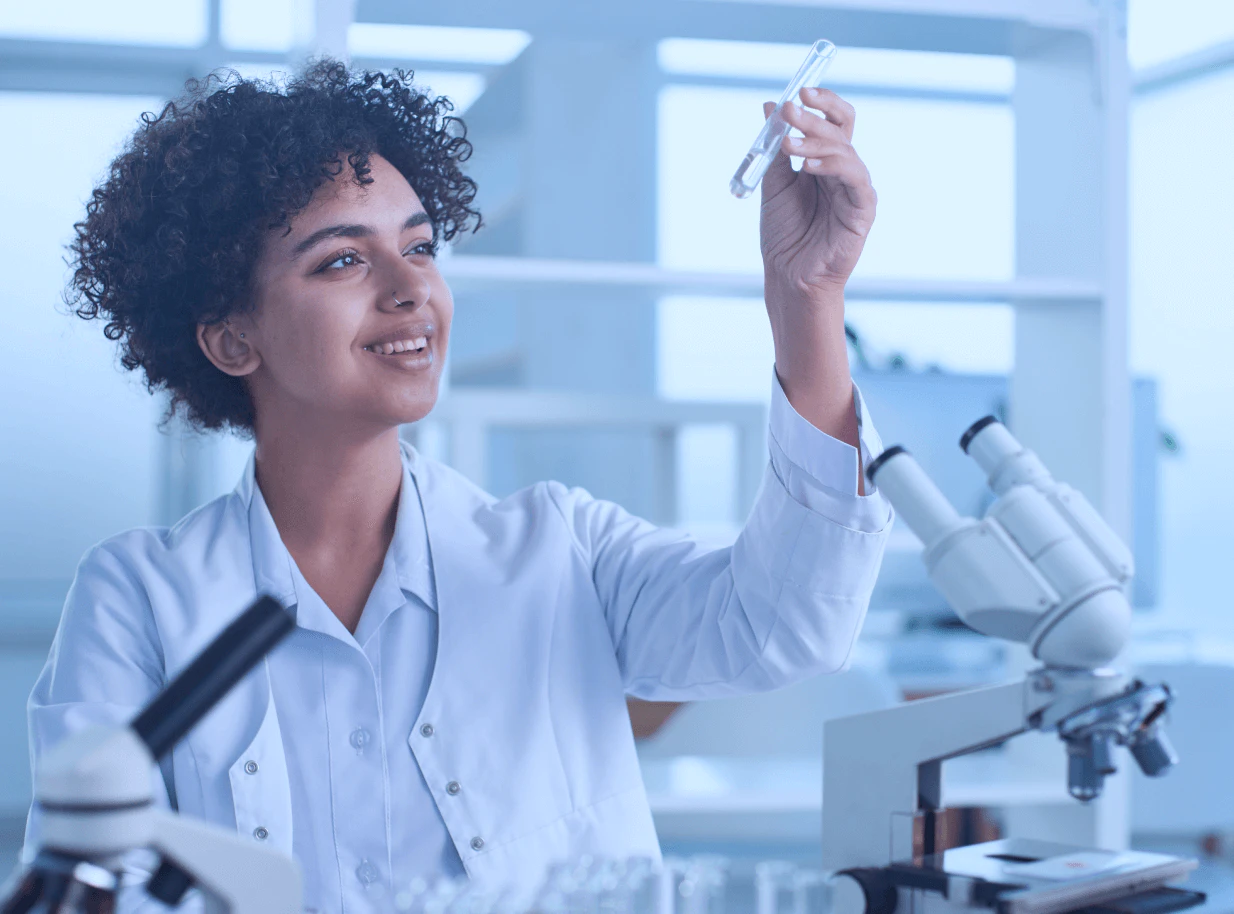 Jovem pesquisadora negra de cabelos cacheados usando jaleco branco segurando tubo de ensaio transparente contra a luz, sorrindo em laboratório moderno com microscópio e equipamentos científicos ao fundo