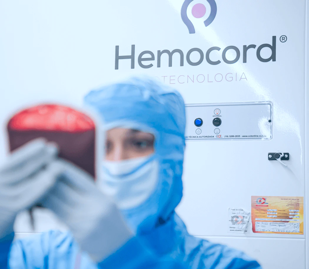 Profissional de saúde usando equipamentos de proteção azuis operando sistema criogênico Hemocord Tecnologia para armazenamento de células-tronco de cordão umbilical, com painel de controle digital e etiquetas de identificação das amostras
