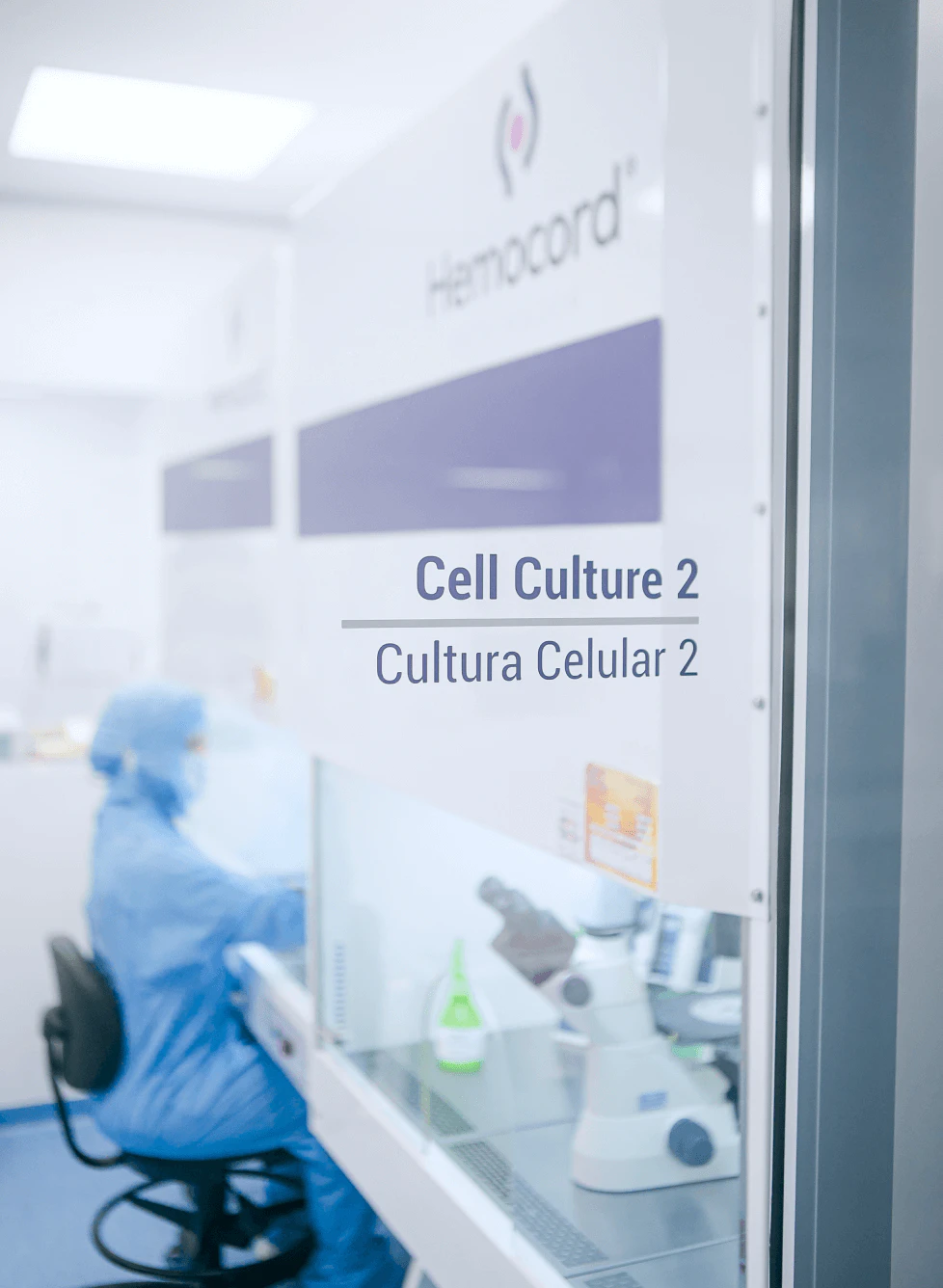 Porta de vidro com placa identificando sala de cultura celular 2, mostrando ambiente controlado com pesquisador e equipamentos laboratoriais ao fundo