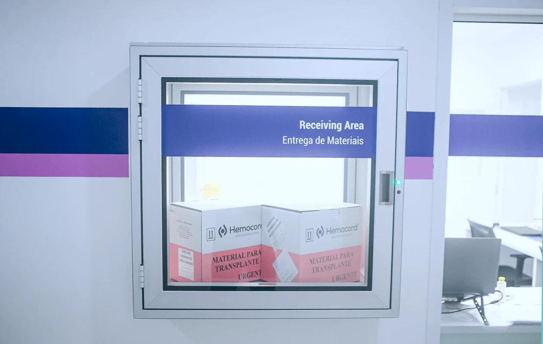 Pass-through hospitalar com compartimento de vidro identificado como "Receiving Area - Entrega de Materiais" contendo caixas térmicas Hemocord etiquetadas como "Material para Transplante Urgente" em ambiente laboratorial com faixas azuis e roxas nas paredes
